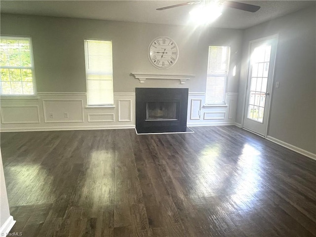 unfurnished living room with wainscoting, a decorative wall, dark wood finished floors, a ceiling fan, and a fireplace with flush hearth