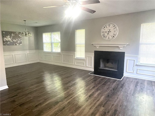 unfurnished living room with ceiling fan with notable chandelier, wainscoting, a decorative wall, and dark wood-style flooring