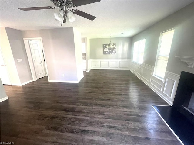 unfurnished living room featuring wainscoting, ceiling fan with notable chandelier, a decorative wall, and dark wood-style floors
