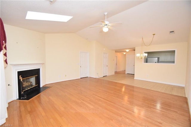 unfurnished living room featuring vaulted ceiling with skylight, visible vents, a glass covered fireplace, wood finished floors, and ceiling fan with notable chandelier