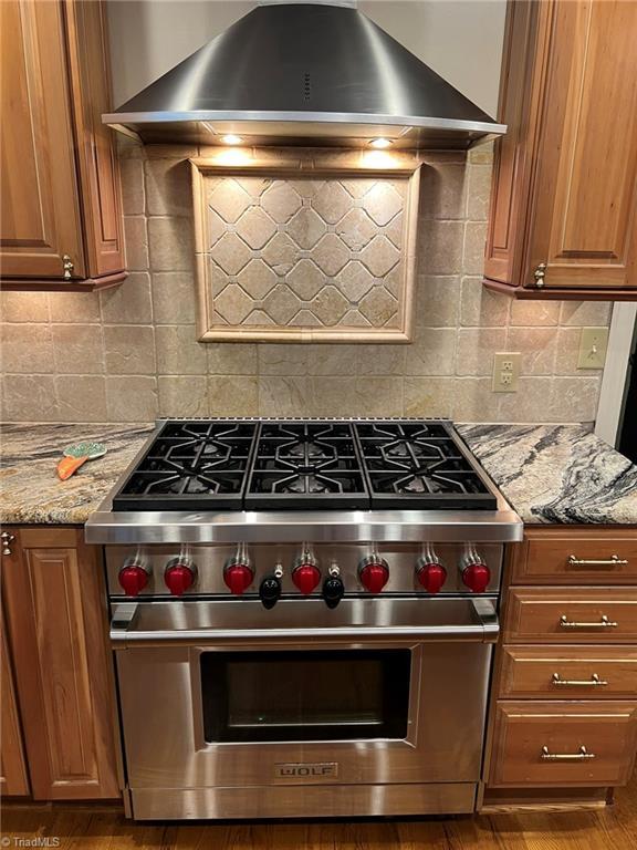 590 Knob View Dr, Winston-Salem, NC 27104 - kitchen with luxury range, wall chimney range hood, brown cabinets, light stone countertops, and decorative backsplash