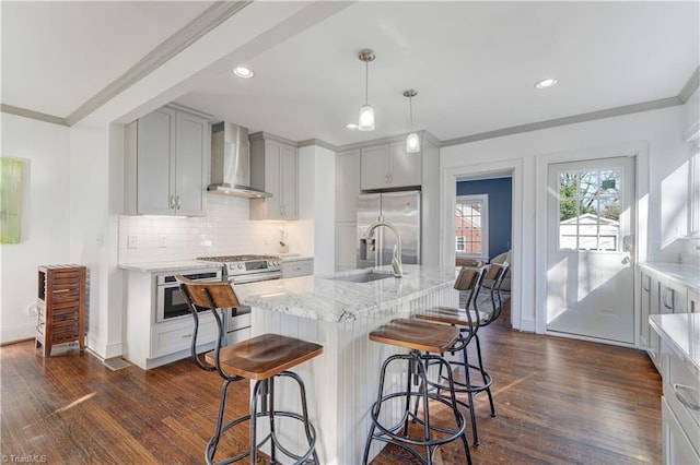 kitchen featuring wall chimney exhaust hood, stainless steel appliances, decorative light fixtures, and an island with sink