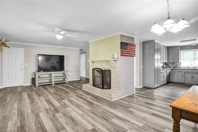 unfurnished living room featuring sink, a brick fireplace, wood-type flooring, ceiling fan with notable chandelier, and ornamental molding