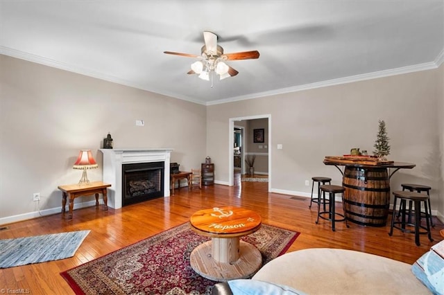 living area with a fireplace, ceiling fan, crown molding, baseboards, and wood finished floors