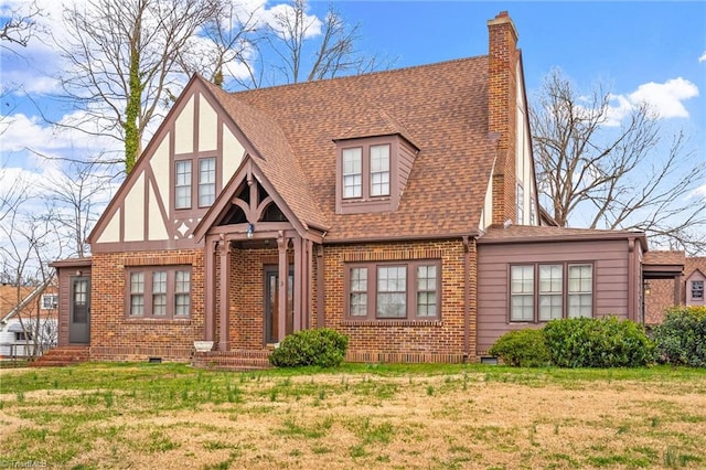 english style home featuring entry steps, brick siding, a chimney, roof with shingles, and a front yard