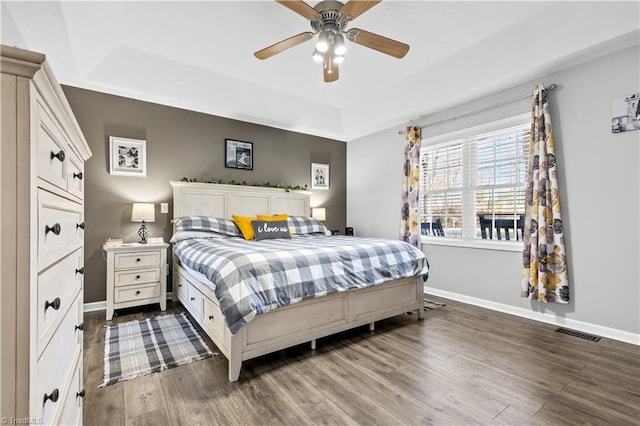 bedroom featuring a tray ceiling, dark wood-style flooring, visible vents, a ceiling fan, and baseboards