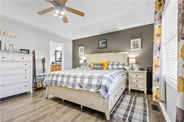 bedroom featuring a tray ceiling, visible vents, ensuite bathroom, a ceiling fan, and wood finished floors