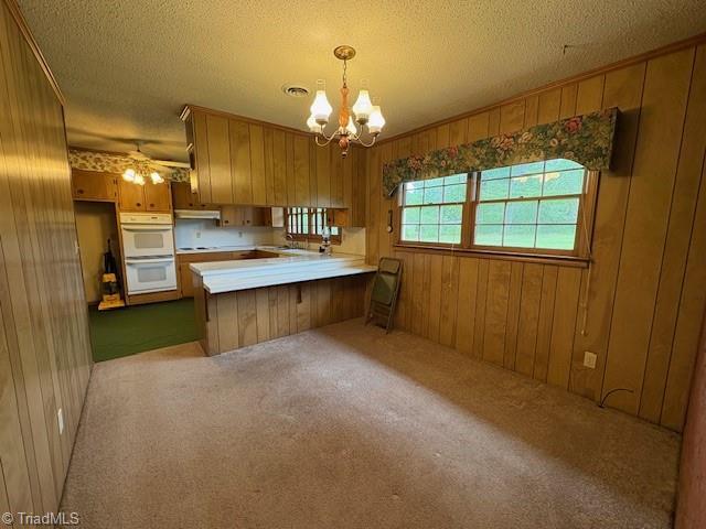 kitchen with a peninsula, brown cabinetry, white double oven, wooden walls, and a chandelier
