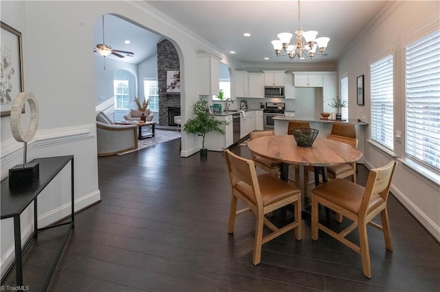 dining area with dark wood-type flooring, a healthy amount of sunlight, ceiling fan with notable chandelier, and a stone fireplace