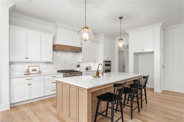 kitchen featuring decorative light fixtures, light wood-style floors, stainless steel double oven, white cabinets, and a kitchen island with sink