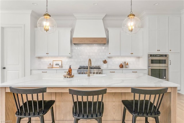 kitchen with light stone counters, a center island with sink, double oven, white cabinetry, and premium range hood