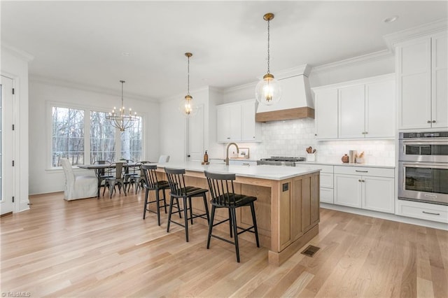 kitchen featuring double oven, white cabinets, light countertops, hanging light fixtures, and an island with sink