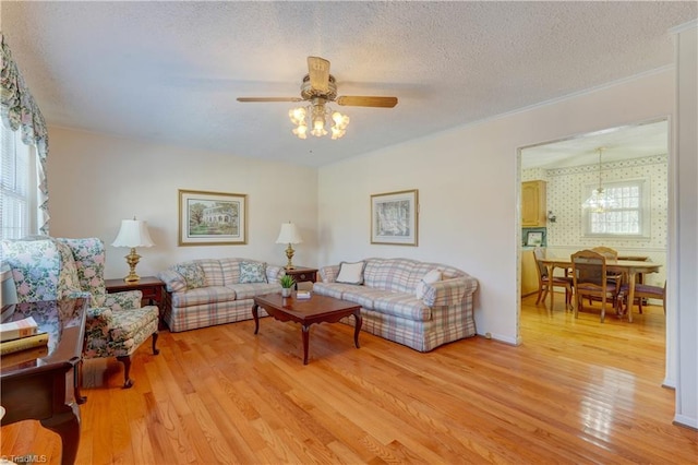 living room with light wood finished floors, wallpapered walls, a ceiling fan, crown molding, and a textured ceiling