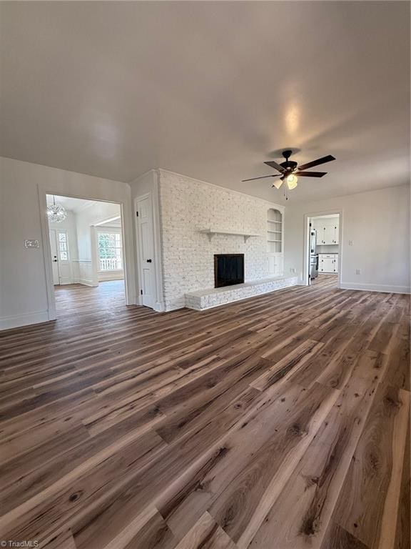 unfurnished living room featuring wood finished floors, a brick fireplace, baseboards, and ceiling fan with notable chandelier