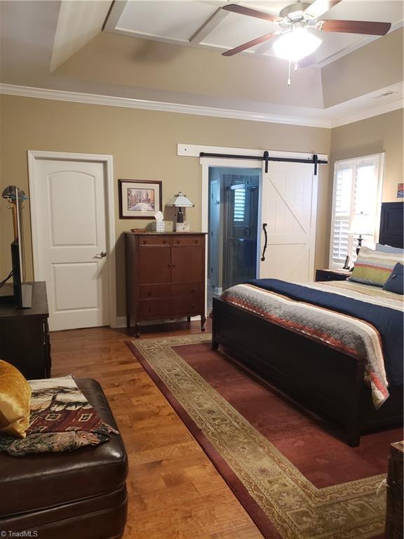 bedroom with ornamental molding, a tray ceiling, hardwood / wood-style flooring, ceiling fan, and a barn door