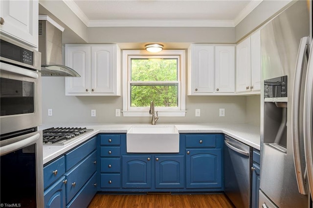 kitchen with blue cabinetry, appliances with stainless steel finishes, wall chimney range hood, ornamental molding, and a sink