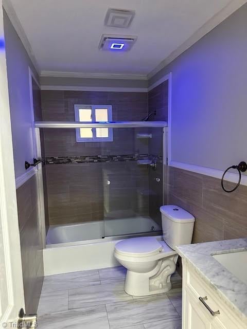bathroom with combined bath / shower with glass door, crown molding, toilet, vanity, and tile walls