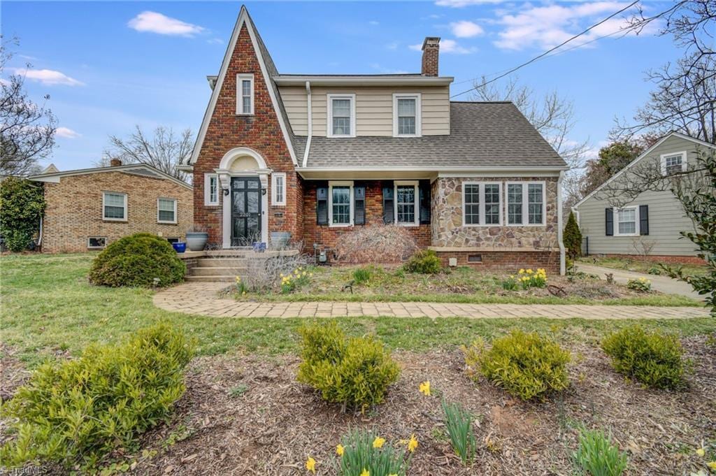 english style home featuring a shingled roof, a front yard, brick siding, and a chimney