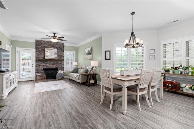 dining room featuring crown molding, a stone fireplace, wood-type flooring, and ceiling fan with notable chandelier