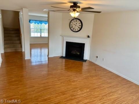 unfurnished living room featuring baseboards, a ceiling fan, a fireplace with flush hearth, wood finished floors, and stairs