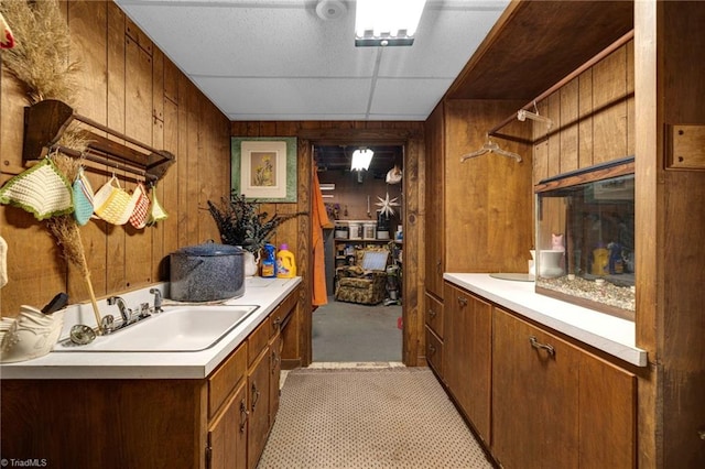 kitchen with a drop ceiling, light colored carpet, wooden walls, and sink