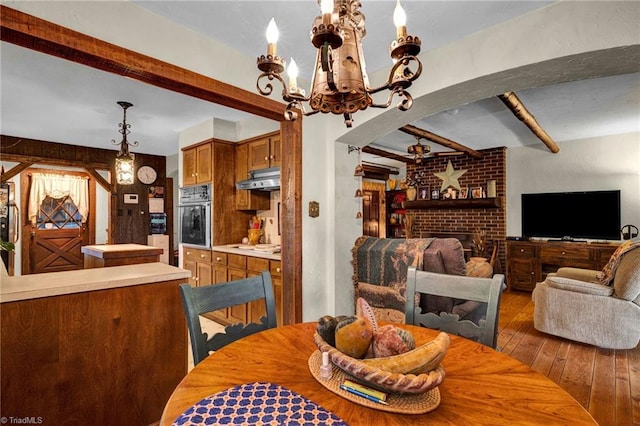 dining area with wood-type flooring, brick wall, an inviting chandelier, beam ceiling, and a brick fireplace