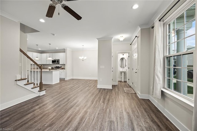 unfurnished living room featuring dark wood-style flooring, ornamental molding, ceiling fan with notable chandelier, and baseboards
