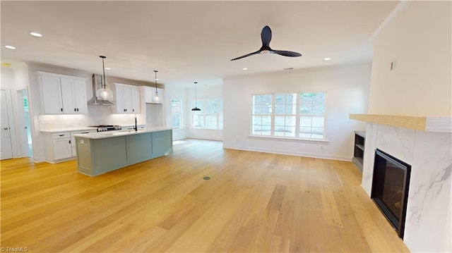 kitchen featuring pendant lighting, wall chimney exhaust hood, light wood-type flooring, an island with sink, and white cabinetry