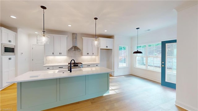 kitchen featuring light countertops, wall chimney range hood, stainless steel microwave, and a center island with sink