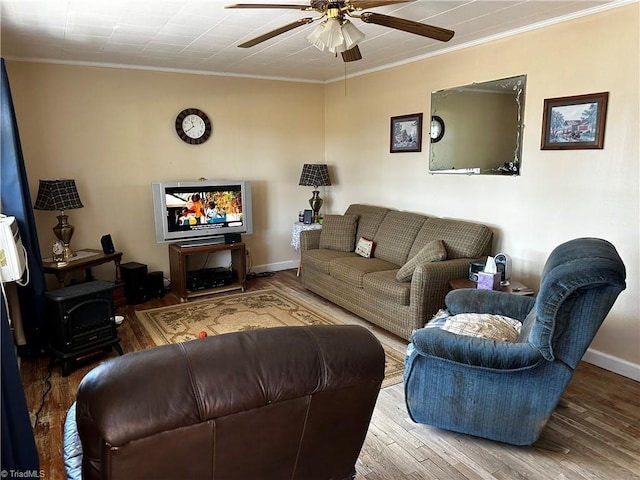 living area with wood finished floors, a ceiling fan, baseboards, ornamental molding, and a wood stove