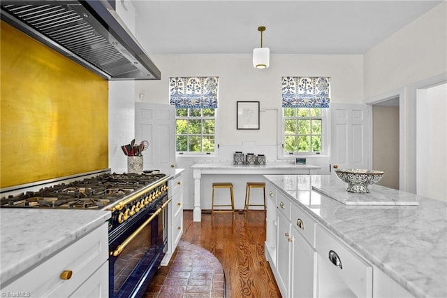 kitchen with range with two ovens, wall chimney range hood, pendant lighting, white cabinets, and light stone countertops