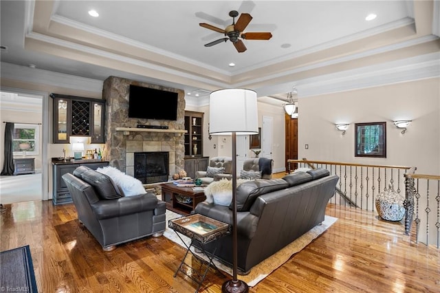 living room with bar, a stone fireplace, wood-type flooring, ornamental molding, and a tray ceiling