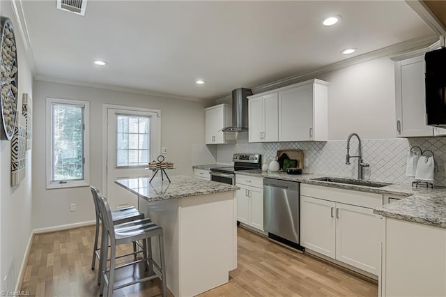 kitchen featuring appliances with stainless steel finishes, a sink, ornamental molding, wall chimney range hood, and light wood finished floors