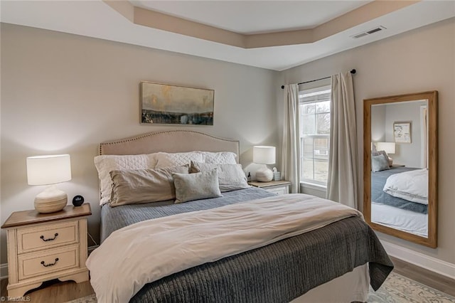 bedroom with a tray ceiling, visible vents, baseboards, and wood finished floors