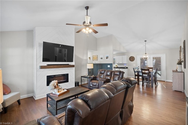 living area featuring a large fireplace, baseboards, lofted ceiling, wood finished floors, and ceiling fan with notable chandelier