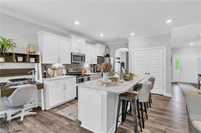 kitchen featuring arched walkways, wood finished floors, an island with sink, appliances with stainless steel finishes, and white cabinetry