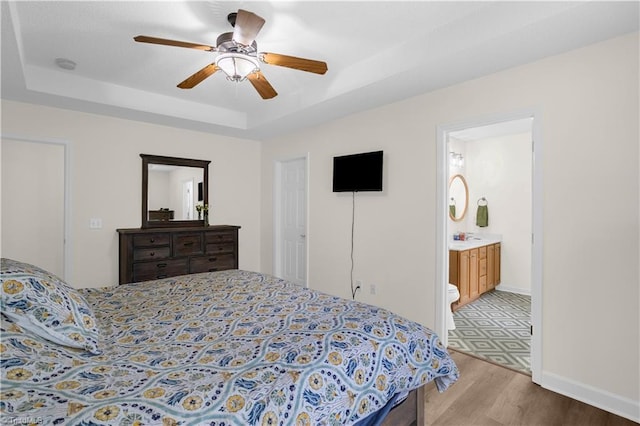 bedroom featuring baseboards, a raised ceiling, a ceiling fan, light wood finished floors, and ensuite bath