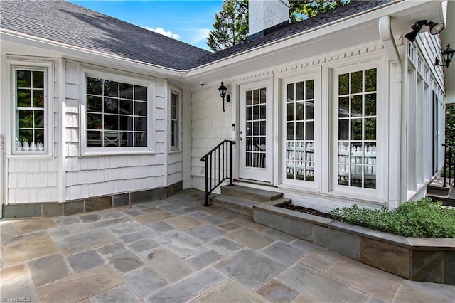 view of exterior entry featuring roof with shingles, a chimney, and a patio