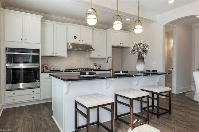 kitchen with under cabinet range hood, dark countertops, backsplash, stainless steel appliances, and arched walkways