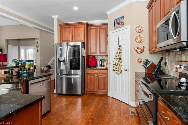 kitchen with appliances with stainless steel finishes, backsplash, dark stone countertops, crown molding, and hardwood / wood-style flooring