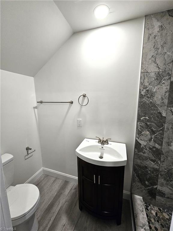 bathroom with toilet, vaulted ceiling, a tile shower, wood finished floors, and vanity