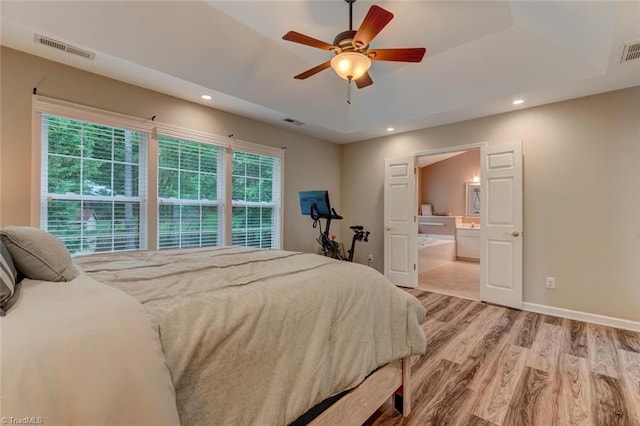 bedroom with light wood-type flooring, a raised ceiling, recessed lighting, ensuite bathroom, and baseboards
