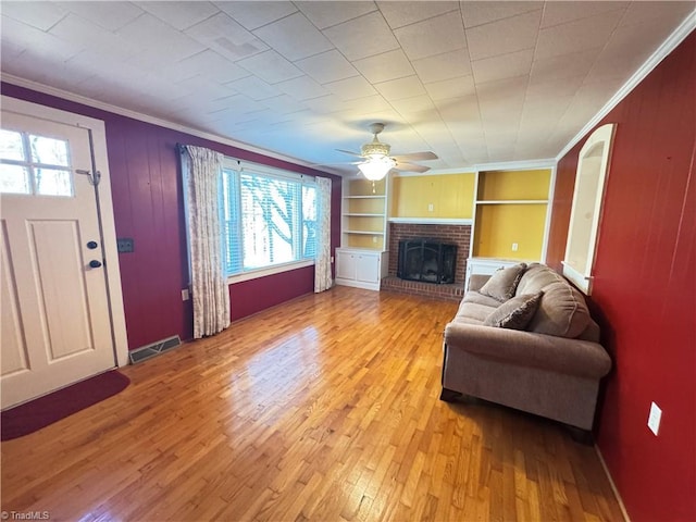 living room with ceiling fan, a fireplace, ornamental molding, built in shelves, and light wood-type flooring