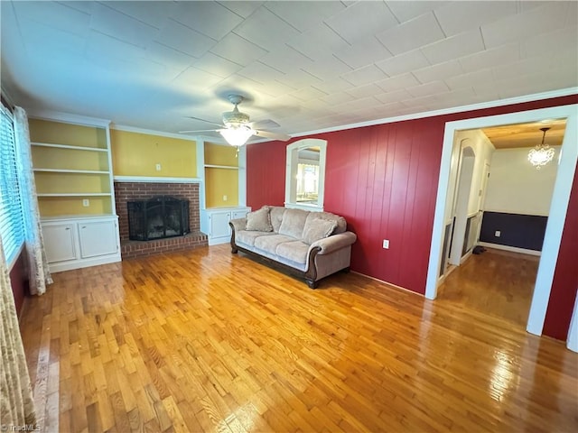 unfurnished living room with crown molding, hardwood / wood-style flooring, built in shelves, a brick fireplace, and ceiling fan with notable chandelier