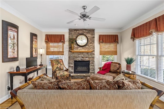 living area with a stone fireplace, wood finished floors, a ceiling fan, baseboards, and crown molding