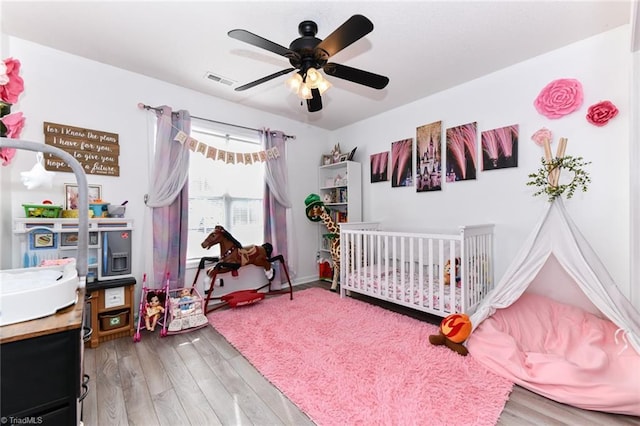 bedroom with visible vents, a nursery area, ceiling fan, and wood finished floors