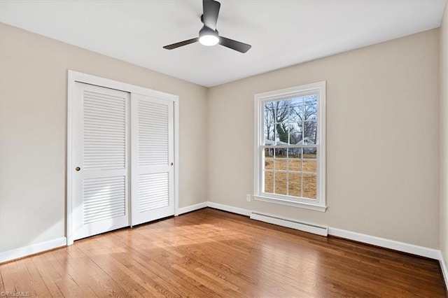 unfurnished bedroom featuring a closet, baseboard heating, ceiling fan, wood finished floors, and baseboards