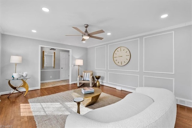 living area with ceiling fan, a decorative wall, wood finished floors, visible vents, and crown molding