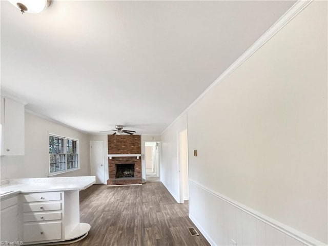 unfurnished living room featuring a ceiling fan, a wainscoted wall, dark wood-style flooring, crown molding, and a fireplace