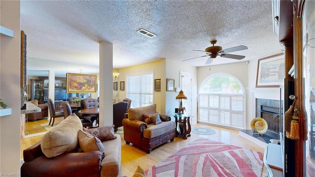 living area with ceiling fan, a fireplace with raised hearth, wood finished floors, a textured ceiling, and ornate columns